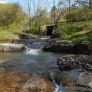 A stream flows under a small bridge in wooded area.