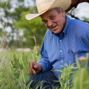 Bill Sproul, a rancher wearing a button-up blue shirt and ivory-colored cowboy hat, crouches in grass with his horse visible in the background