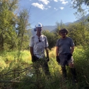 A tall man in hardhat, dirty t-shirt, and ear protection stands next to a shorter man in straw hat, shorts, and t-shirt, both wearing work gloves, in a field.