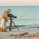 A man using a large video camera standing on a beach filming a crab in the water