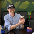 A smiling, bearded man in a ballcap holds up a small carved salmon