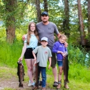 A man with two girls and a boy stand in front of trees, all 3 kids holding huge trout.