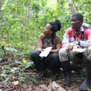 Two people dressed in field clothes take a break in the forest, holding notebooks. The woman, sitting to the left, looks up towards the canopy