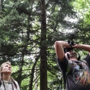 Two people standing in a forest looking up, one using binoculars