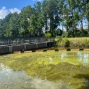 A concrete bridge structure with vertical grates along the sides extending into the water. In the foreground, a floating boom reaches diagonally across the canal.