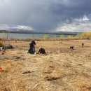 People scattered across a brown hillside, kneeling and standing, with the lake, yellow trees, and dramatic clouds visible behind.