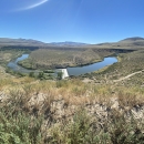 A river running through a desert sage landscape with small mountains in the backdrop and bright blue skies above.