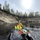 Tim Ericson, sitting at the front of a boat, paddles on the Deshka River. The photo is taken from the back of the boat, and Ericson looks back at the camera over his shoulder. The boat is filled with supplies, including moose antlers. 