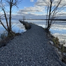 a stone jetty with sparse trees cuts through a still lake that reflects blue skies and clouds