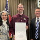 Three people smile standing in front of an American flag, and the man in the middle holds an award