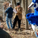 Two men, one holding a guitar, sing next to a blue goose mascot.