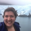 Woman with brown hair and blue jacket in front of water and a white and light blue glacier in the background