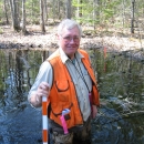 A man holding a yard stick and wearing an orange vest and waders standing in a shallow pond in a forested area