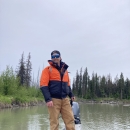 A person wearing a float coat and work pants stands on a boat parked on a river.