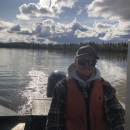 A person wearing an orange lifejacket, flannel shirt, and hooded sweater, along with a hat and sunglasses,smiles as they steer a boat's rear engine on a river under a partly cloudy sky.