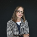 A woman with brown hair and glasses stands in front of a black backdrop smiling at the camera.