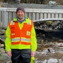 A person wearing an orange visibility vest smiles in front of a bridge with a stream running under it.