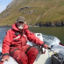 Man wearing orange float suit steers a skiff with water and island in the background.