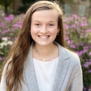 Brynn Garner smiles, standing in front of bushes with pink flowers