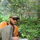 Fish Biologist, Brian Davis, collecting samples in the field. Brian is wearing an orange shirt, tan pants, tan hart and black sunglasses. He is reclining on the ground in a temperate rainforest setting with sampling equipment in front of him. 