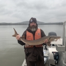 Man holds up a large brown fish with both hands while standing on a boat with a water and hilly backdrop.