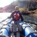 A person in a life vest sits in an inflatable boat holding oars. The boat is is the middle of river surrounded by forests.