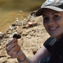 Sitting woman smiling and holding a mussel in front of a creek.