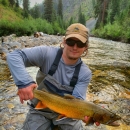 A man in waders kneeling in a stream holds a large fish in his hands. 