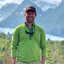 A man standing outside with a field, a glacier, and mountains in the background