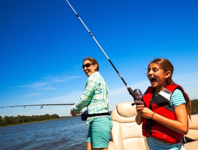 A young girl and her mother laugh as they enjoy fishing on a boat in the middle of a lake in the summer.