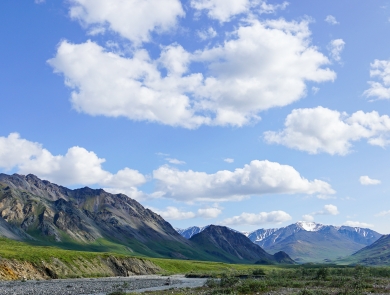 Landscape photo of green mountains, a river bank, and blue sky with clouds