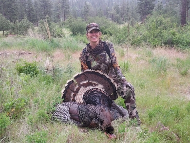 A woman wearing camouflage kneels beneath a turkey she harvested