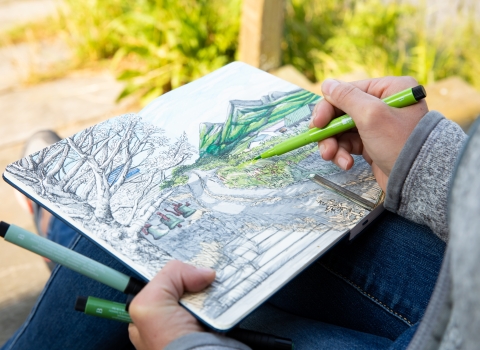 A hand holding a green felt tipped pen coloring in an illustration in a sketchbook of a mountainous scene