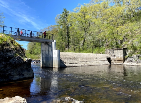 A dam crosses a river, but a portion has been removed to that water can flow through. People stand on an overlook that crosses the open section of the river. 