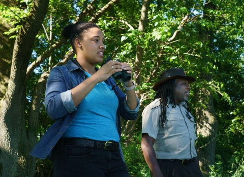 A woman and a man, both U.S. Fish and Wildlife Service employees, look for birds in the woods. The woman holds binoculars.