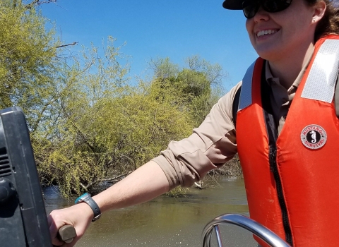 A woman drives a boat wearing a life vest and Fish and Wildlife Service hat