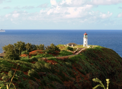 A lighthouse stands on the edge of a peninsular cliff with a large ship in the s