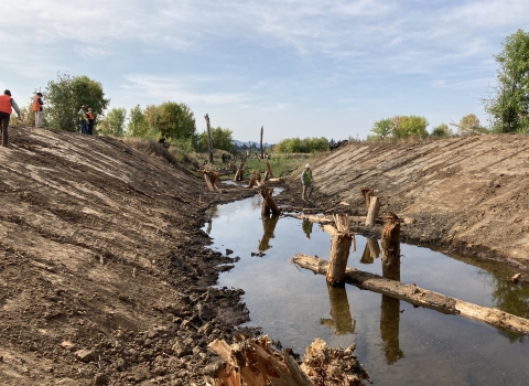 Channel filled with low level of water and large woody debris; construction works at the top of the banks; trees in the distance, with cloudy and blue skies