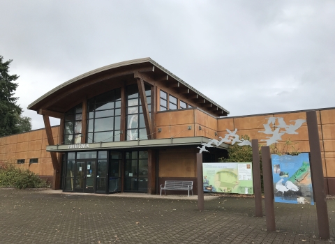 Large building with glass windows and tan siding; angled roof; sign that says "visitor center"; interpretive panels near building; stone paved ground