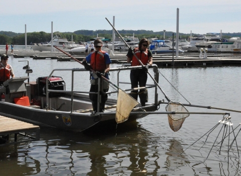 People in brown uniforms and orange flotation gear stand on a boat in a waterway holding nets.