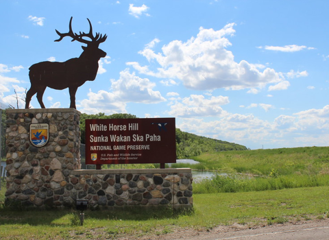 A sign bearing the USFWS logo and a metal cutout of an elk. The sign reads White Horse Hill Sunka Wakan Ska Paha National Game Preserve, United States Fish and Wildlife Service