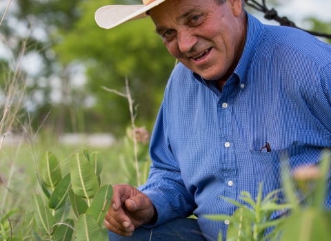 Bill Sproul, a rancher wearing a button-up blue shirt and ivory-colored cowboy hat, crouches in grass with his horse visible in the background