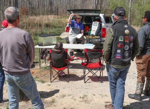 Several adults in a semi-circle, listening to one person sitting on a truck tailgate