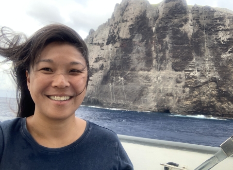 Jiny Kim, wearing a navy blue shirt, stands on a boat with Nihoa in the background.