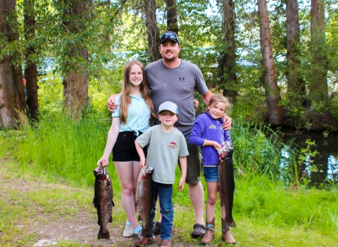 A man with two girls and a boy stand in front of trees, all 3 kids holding huge trout.