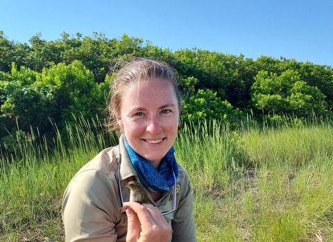 fish and wildlife biologist sits in the marsh holding a sparrow 