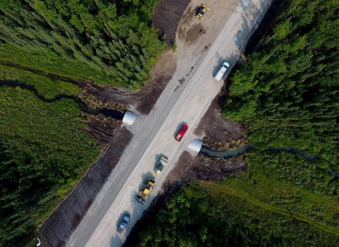 An aerial view of newly replaced culvert on a dirt road in rural Alaska. Cars and construction machines are parked on the road above the culvert, and a stream of water cuts through the greenery of pine trees and foliage.