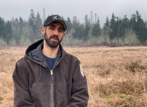 A man in brown jacket a baseball hat stands in a field