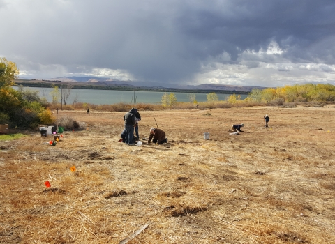 People scattered across a brown hillside, kneeling and standing, with the lake, yellow trees, and dramatic clouds visible behind.