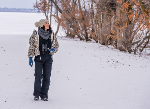 A woman wearing a warm jacket, hat, gloves, and binoculars walks on a snow covered path looking into the trees.